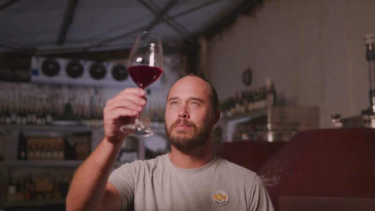 A man with a beard holds up a glass of Boschkloof Epilogue Syrah, inspecting it closely in a dimly lit room with Boschkloof wine bottles and barrels in the background.