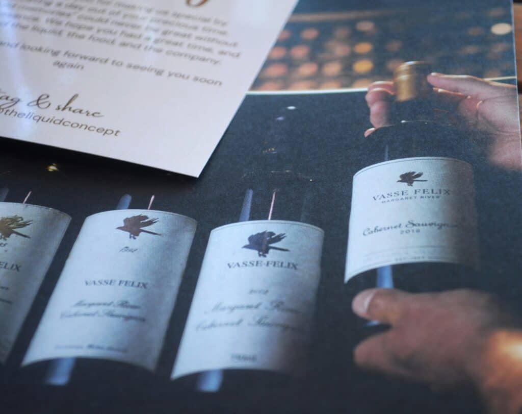 A hand holds a Vasse Felix Cabernet Sauvignon wine bottle next to three similar bottles on a counter, with a partially visible menu or card above them—highlighting how critics can give the same wine a different score.